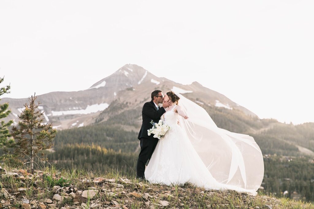 A stunning wedding moment in Big Sky, Montana, featuring a newlywed couple sharing a romantic kiss with the breathtaking Lone Peak mountain in the background. The bride’s veil flows in the wind, adding an ethereal touch to this scenic outdoor wedding portrait.