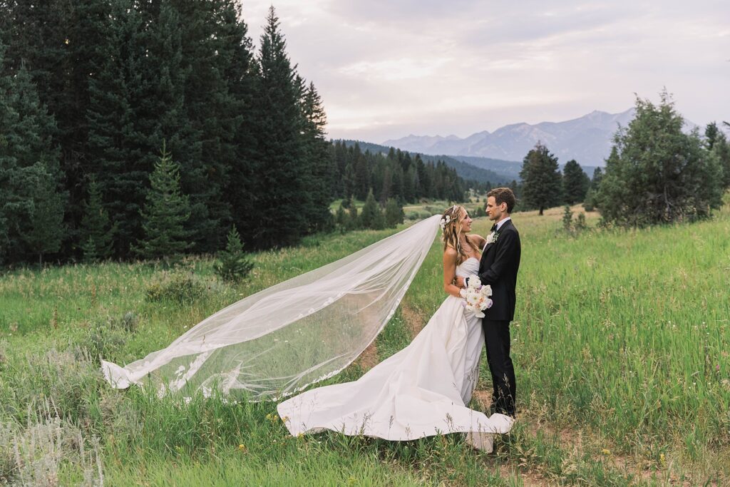 A bride and groom embrace in a lush green meadow at Rainbow Ranch Lodge, with a stunning mountain range in the background. The bride’s veil flows in the wind.