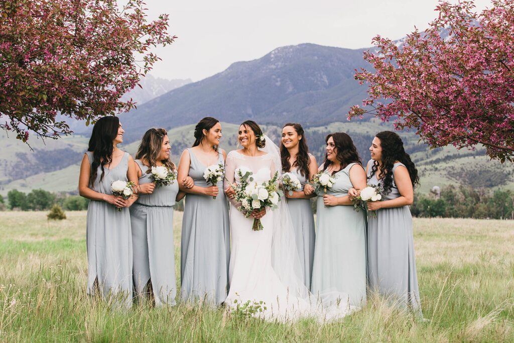 Bride and bridesmaids in soft blue dresses stand beneath blooming trees with mountain views in the background at Yellowstone House in Montana.