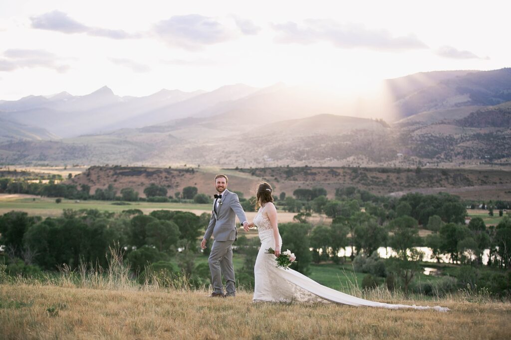 Bride and groom walking hand in hand at Sage Lodge during golden hour, with the Montana mountains glowing in the background.