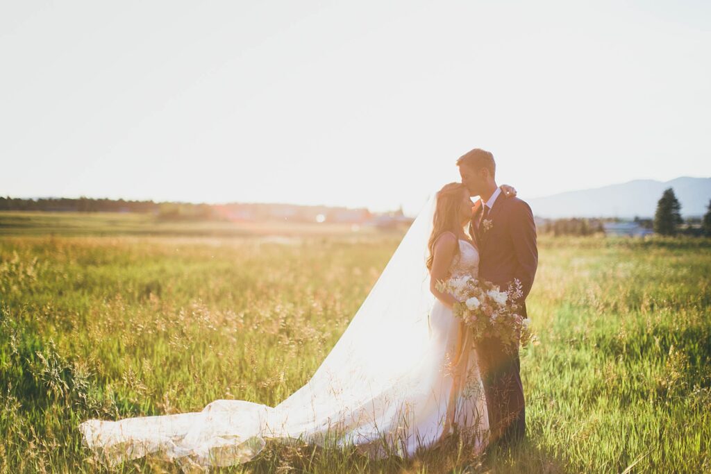 A bride and groom share a romantic embrace in a sunlit field at golden hour, with a flowing veil and lush countryside backdrop at Foster Creek Farm in Belgrade, Montana.