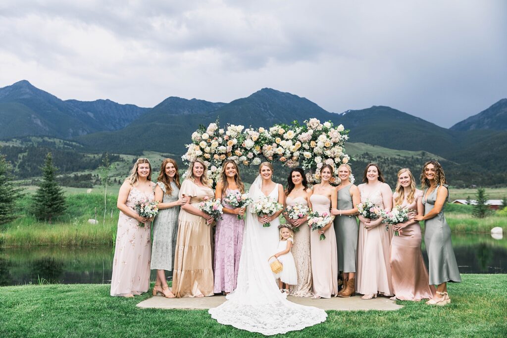 A bride stands with her bridesmaids in front of a floral arch with mountain views at Copper Rose Ranch.